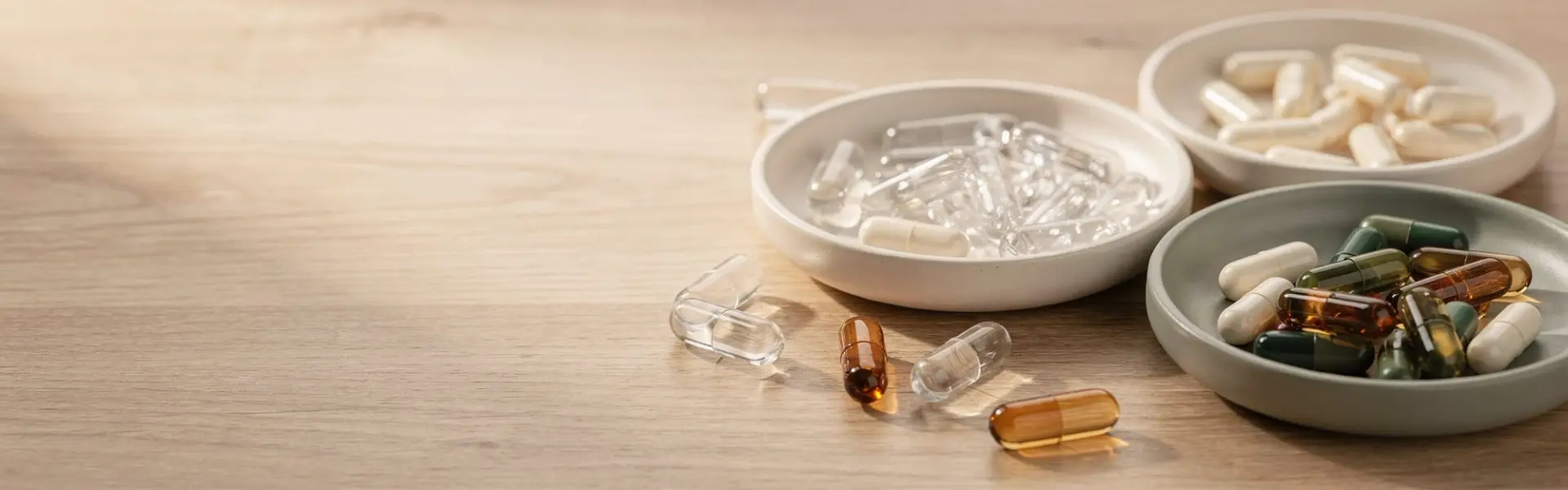 Assortment of transparent, ivory and coloured empty hard capsules in shallow ceramic dishes on a wooden surface